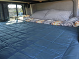 A comfy rv rental bed with blue comforter & pillows in a van, ready for travel. Outside view from window.