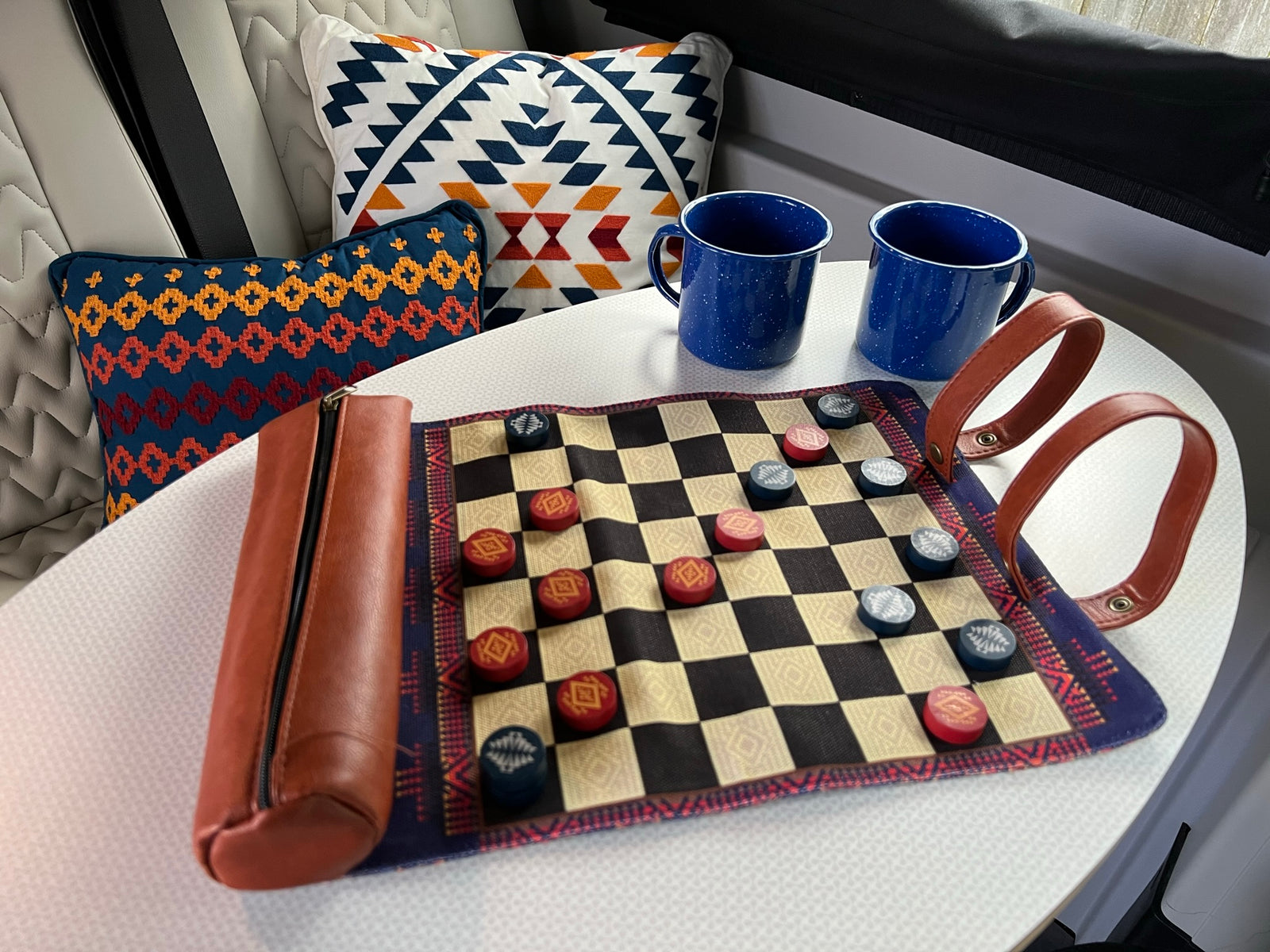 Cozy rv rental setup: a checkers game board, two blue enamel mugs, and patterned pillows. Ideal for relaxing travel.