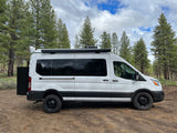 White Ford Transit rv rental camper van parked in a forest with pine trees under a cloudy sky, ready for adventure.