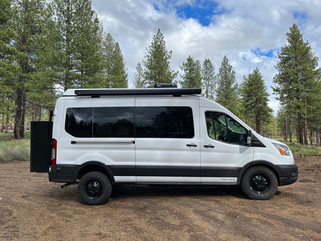 White Ford Transit rv rental camper van parked in a forest with pine trees under a cloudy sky, ready for adventure.