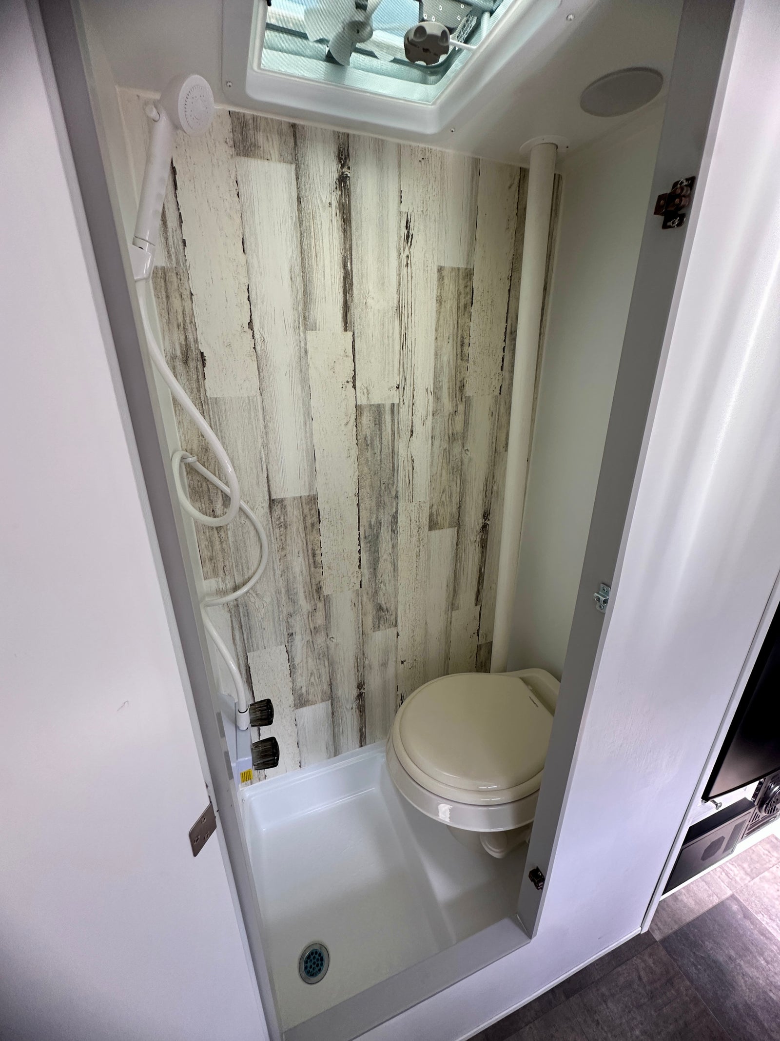 RV rental compact wet bath featuring a toilet, handheld shower, and drain. Light wood-look walls and ceiling vent visible.