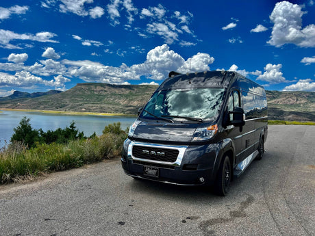 Black RV rental parked by a scenic lake with mountains under a bright blue sky and fluffy clouds.