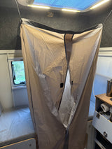 Inside an RV rental, a hanging beige privacy curtain divides the compact interior, revealing a bed, window, and small kitchenette.