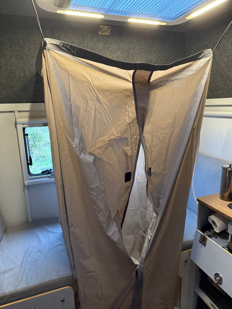 Inside an RV rental, a hanging beige privacy curtain divides the compact interior, revealing a bed, window, and small kitchenette.