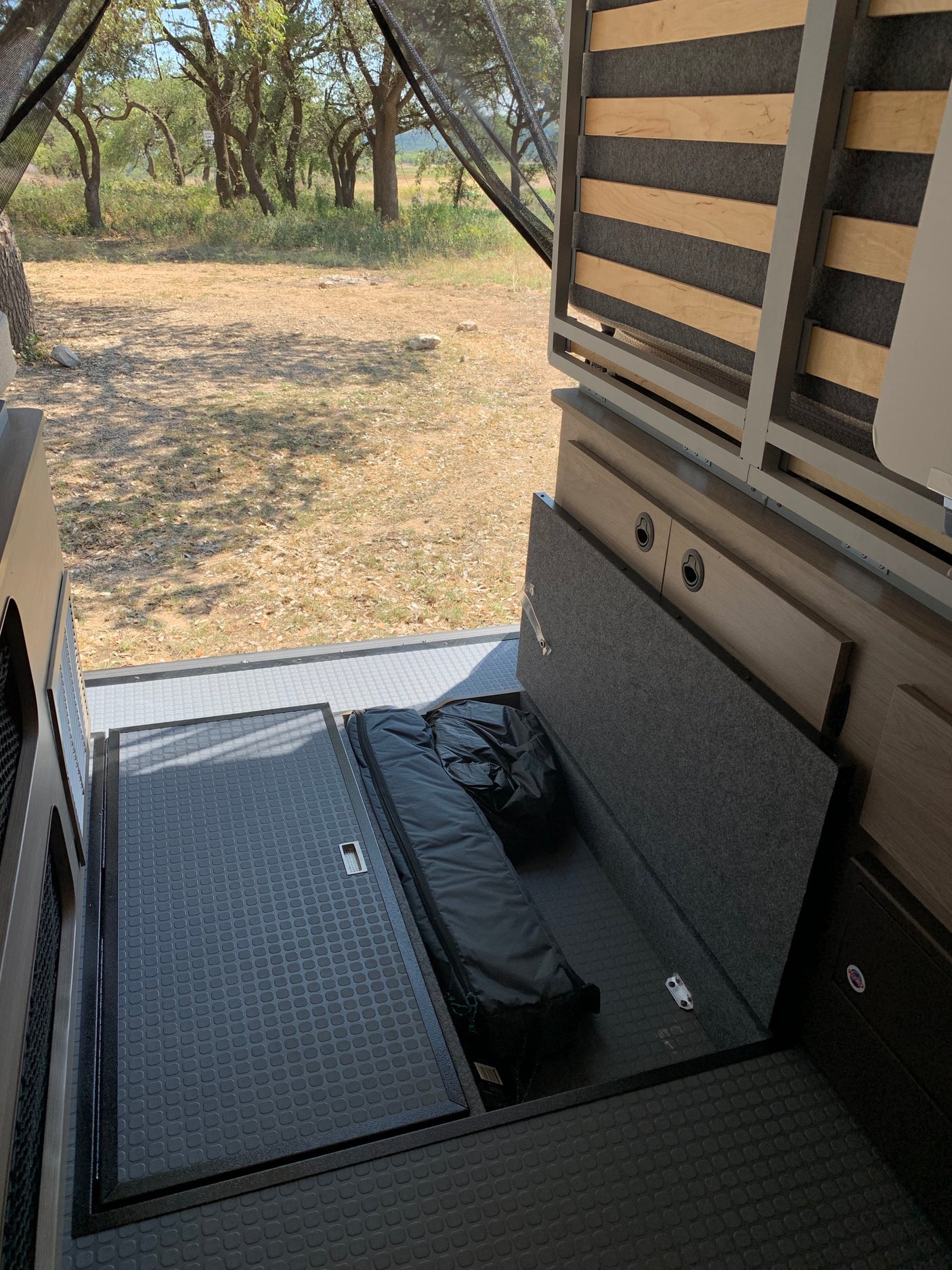 RV rental interior with open storage revealing a sleeping bag & items, bed frame, and outdoor view to trees.