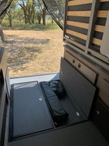 RV rental interior with open storage revealing a sleeping bag & items, bed frame, and outdoor view to trees.