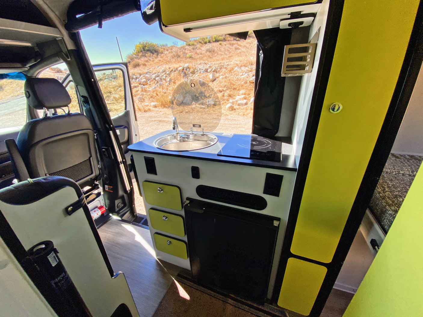Inside a modern rv rental camper van: kitchen with sink, stove, fridge, and bright green accents. View of dry landscape.