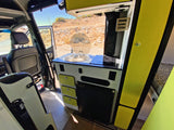 Inside a modern rv rental camper van: kitchen with sink, stove, fridge, and bright green accents. View of dry landscape.
