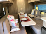 An inviting RV rental interior: banquette dining, coffee mugs on tables, and two plush animals enjoying the serene ocean view.
