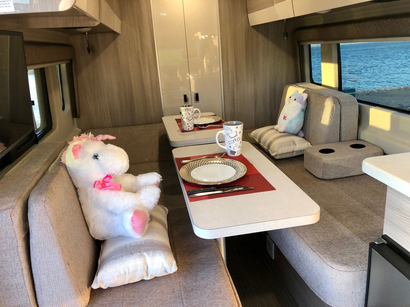 An inviting RV rental interior: banquette dining, coffee mugs on tables, and two plush animals enjoying the serene ocean view. — photo 13