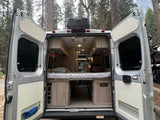 Rear view of an RV rental camper van, doors open to reveal a comfortable bed, light wood cabinetry, and integrated controls.