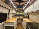 Interior of a stylish rv rental featuring a cozy bed, functional kitchen with sink & stove, and a chic wood-slat ceiling with LED lights.