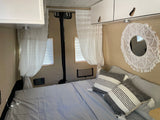 Cozy RV rental bedroom features a light blue bed, white overhead cabinets, macrame mirror, and sheer curtains.