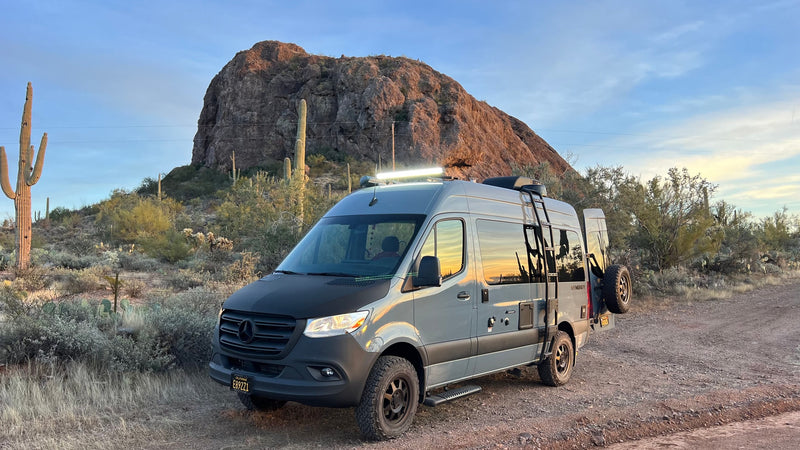  surrounded by desert vegetation and towering rock formations.|Grey Mercedes-Benz Sprinter RV rental parked in a desert landscape with saguaro cacti and a rocky mountain.|Explore stunning landscapes with an rv rental! Grey camper van near a towering desert rock formation under a bright blue sky.|Grey Mercedes Sprinter rv rental van with side door open — photo 4