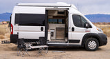 White RV rental camper van parked in desert with side door open to interior kitchen. Folding chairs/table outside.