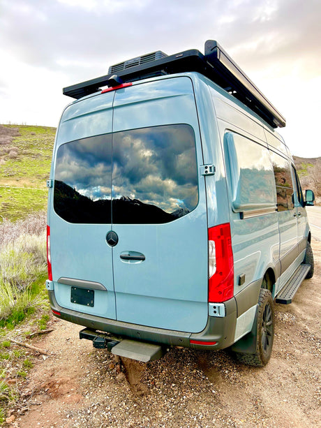 Rear view of a light blue Mercedes Sprinter campervan, ideal for RV rental. Features roof rack, side steps, and mountain reflections.