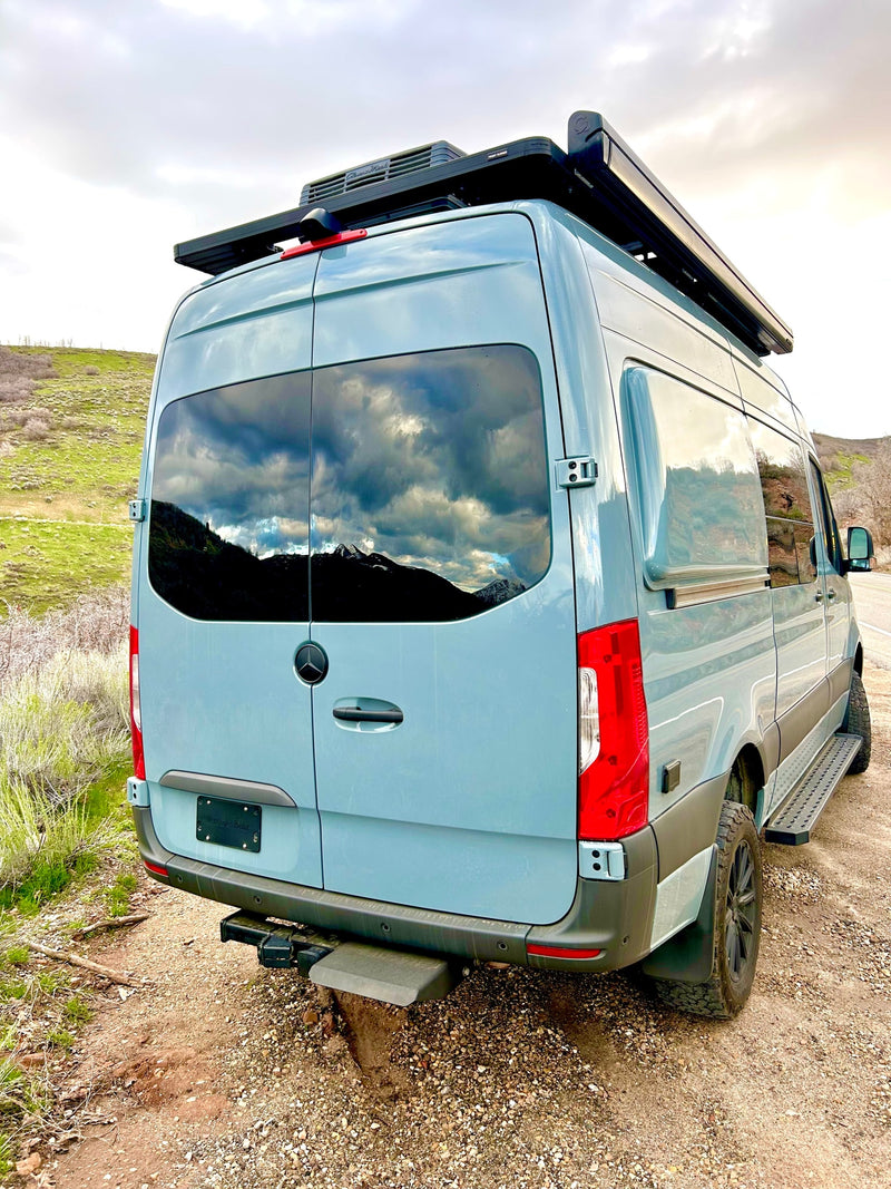 Rear view of a light blue Mercedes Sprinter campervan, ideal for RV rental. Features roof rack, side steps, and mountain reflections. — photo 9