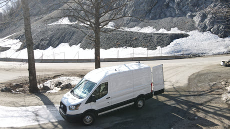 White van, like an RV rental, parked by a snowy mountain road with bare trees. Rear doors are open.
