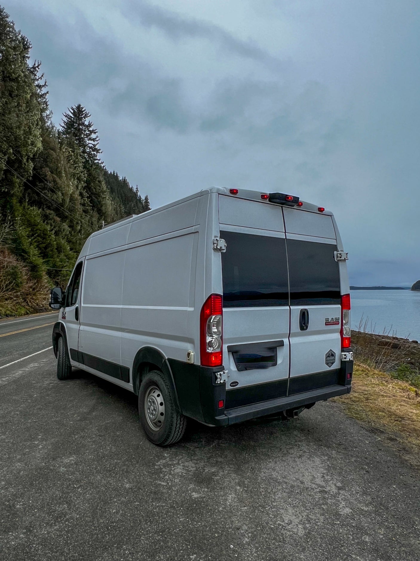 Light gray camper van, an RV rental, parked on a scenic coastal road. Perfect for nature exploration under cloudy skies.