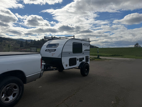Sunray Sport RV rental trailer hitched to a truck, parked on asphalt near a lake, surrounded by green hills under a cloudy sky.