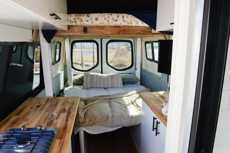 Inside a cozy rv rental camper van. Features a comfortable bed, kitchen counter with stove, and overhead storage.