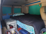 RV rental interior: a comfy bed with tropical sheets, teal window covers, and a large cooler.