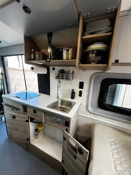 Compact RV rental kitchen: sink, stovetop, and storage cabinets. Doors are open, showing dishes and cleaning supplies.