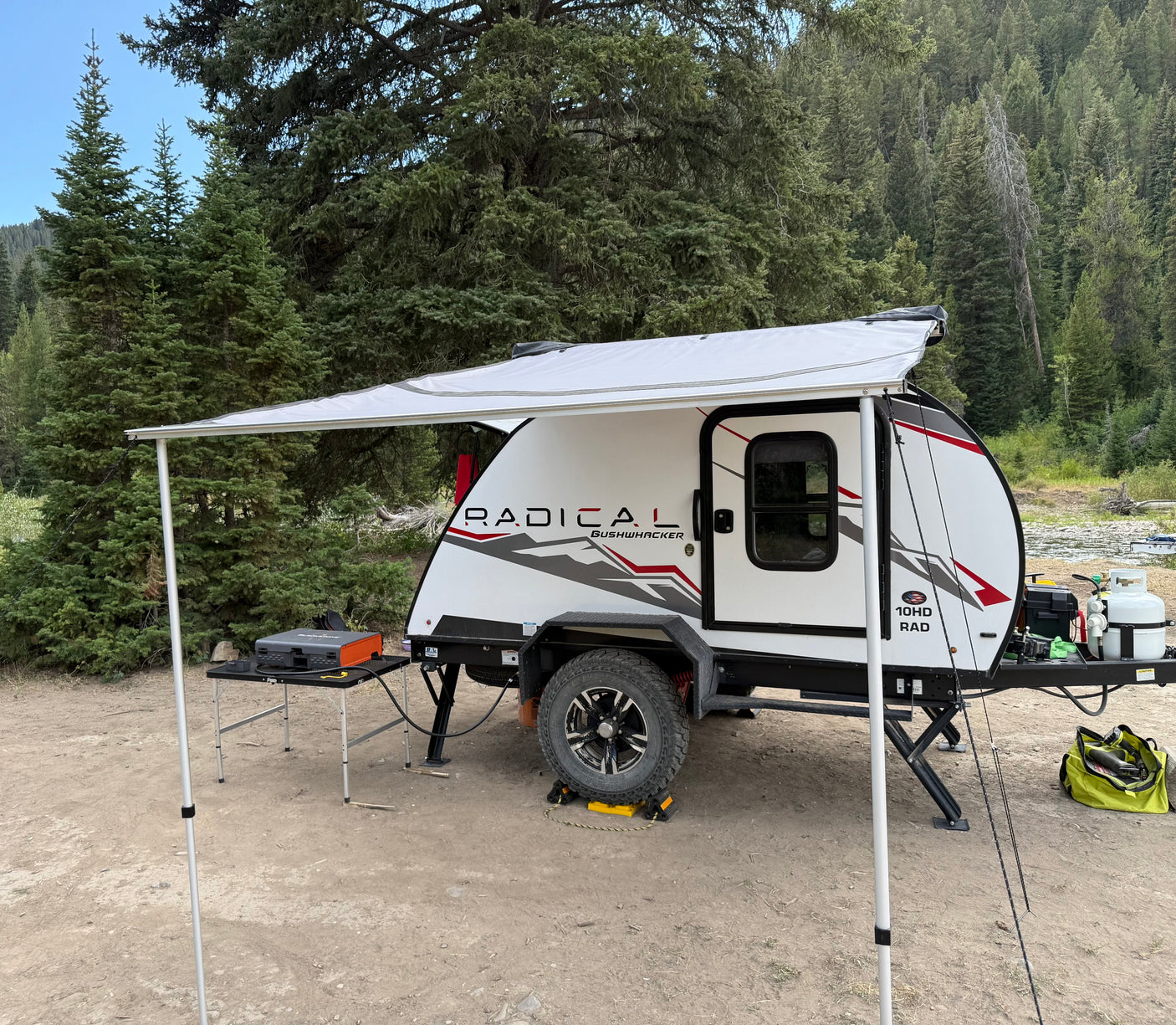 An RV rental, a compact teardrop trailer, is set up with an awning for comfortable camping in a peaceful forest campsite.