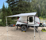 An RV rental, a compact teardrop trailer, is set up with an awning for comfortable camping in a peaceful forest campsite.