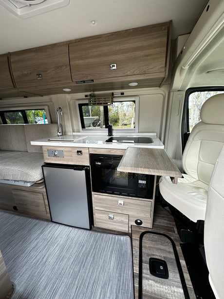 Inside an RV rental: a bright, compact living area featuring a well-equipped kitchenette with microwave, sink, and refrigerator.