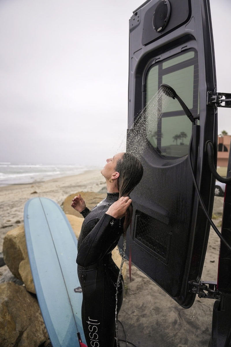 Woman in wetsuit takes an RV rental's outdoor shower on beach near her surfboard. Perfect for surfing post-rinse. — photo 7