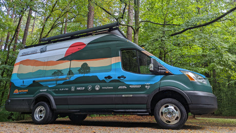 Adventure-ready **RV rental** van with a scenic mural & sponsor logos, parked amidst green trees.