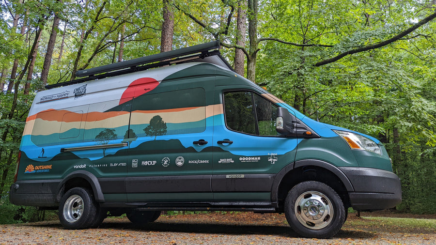 Adventure-ready **RV rental** van with a scenic mural & sponsor logos, parked amidst green trees.