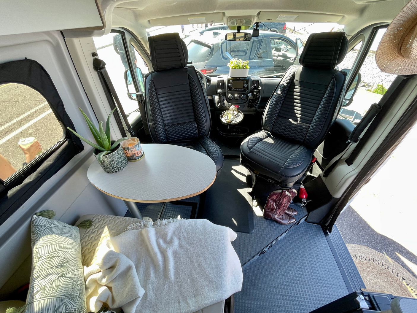 Inside a bright RV rental: black quilted front seats, a round dining table, and a cozy lounge area with pillows and a blanket.