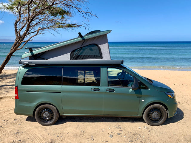 Green pop-top camper van parked on a sandy beach by the clear blue ocean. Ideal for your next RV rental trip.
