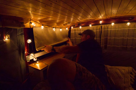 Enjoy a cozy rv rental experience! Man adjusts window shade inside a warmly lit camper van with string lights.