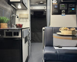 Inside an RV rental: a compact living space with kitchen, sink, fridge, dinette, and control panel on the right.