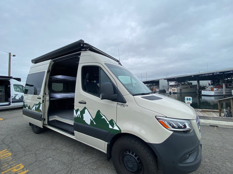 This custom Mammoth adventure van, an RV rental, with bunk beds inside, is parked by boats at a marina.