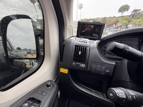 RV rental dashboard view: Interior controls and monitor, while the side mirror reflects a beach scene under an overcast sky.