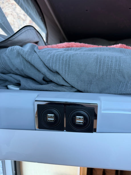 RV rental interior: dual USB charging ports on a white panel, with soft blankets and a mesh window visible in the background.