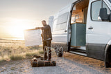 Man assembles a camp table next to his white RV rental van, showcasing its open modern interior and outdoor setup at sunset.
