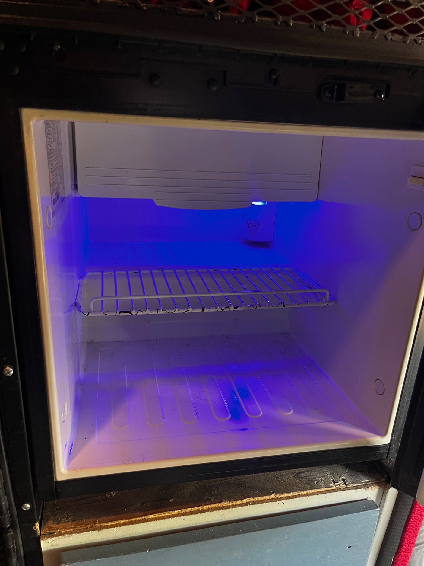 Empty RV rental mini-fridge interior glows with a blue light, featuring a wire shelf, ready for groceries.