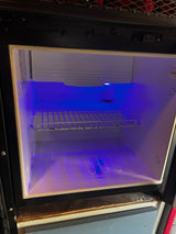 Empty RV rental mini-fridge interior glows with a blue light, featuring a wire shelf, ready for groceries.