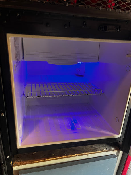 Empty RV rental mini-fridge interior glows with a blue light, featuring a wire shelf, ready for groceries.