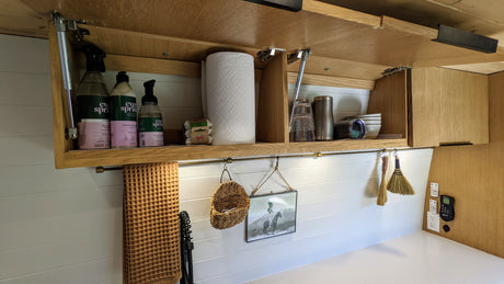 A tidy rv rental kitchen featuring an open overhead cabinet with essentials, hanging decor, and organized counter space.