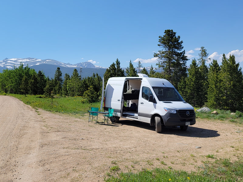 White RV rental camper van on a dirt road, open door, with chairs and table, overlooking snow-capped mountains and pines. — photo 7