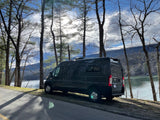 A dark gray camper van, ideal for an RV rental, is parked by a serene lake framed by tall trees and distant mountains.