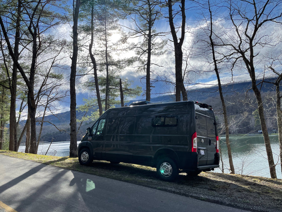 A dark gray camper van, ideal for an RV rental, is parked by a serene lake framed by tall trees and distant mountains.