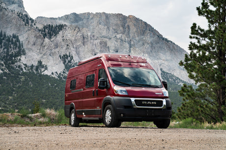 Red camper van for RV rental, parked on a mountain dirt road, surrounded by nature and evergreen trees.