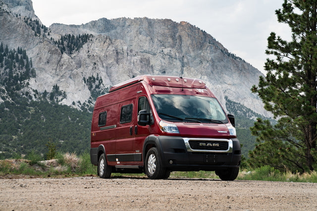 Red camper van for RV rental, parked on a mountain dirt road, surrounded by nature and evergreen trees.